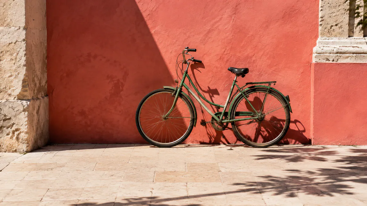 A weathered jade-green bicycle rests against a coral courtyard wall in strong midday light.