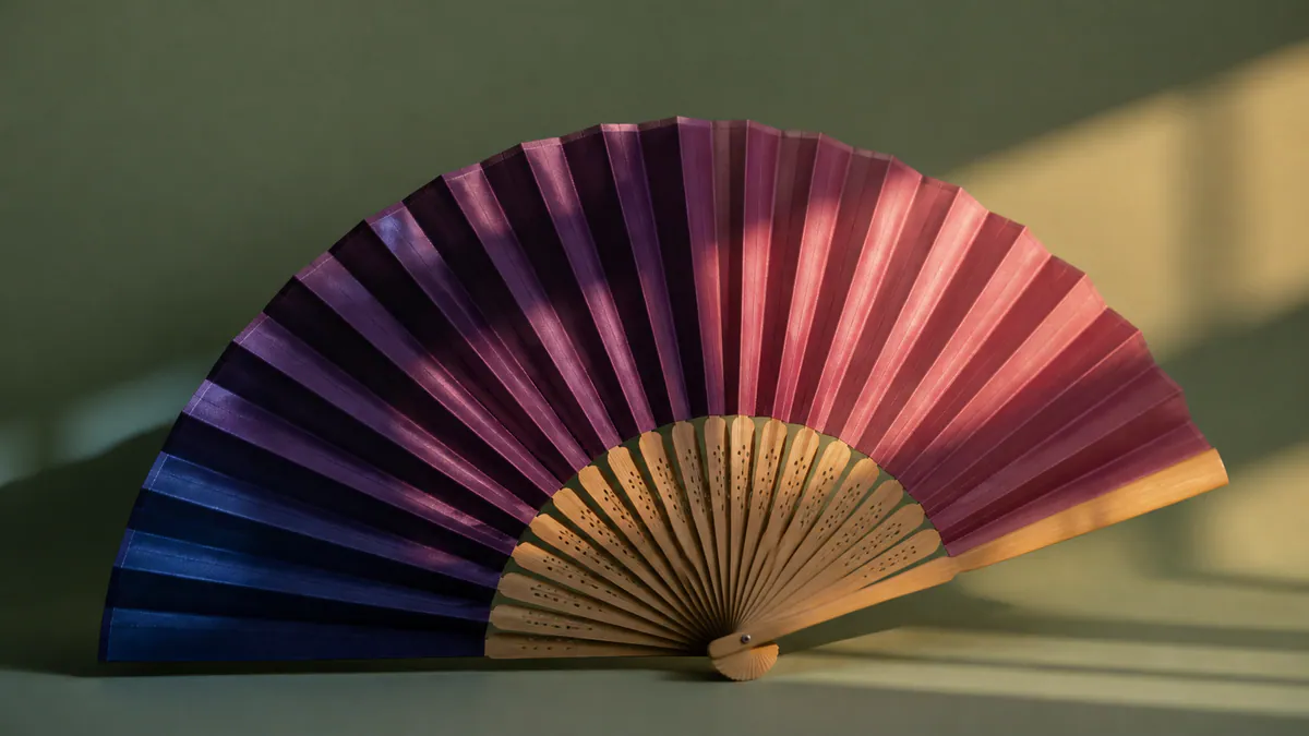 A deep plum and coral pleated fan rests in amber and indigo light on a matte sage surface.